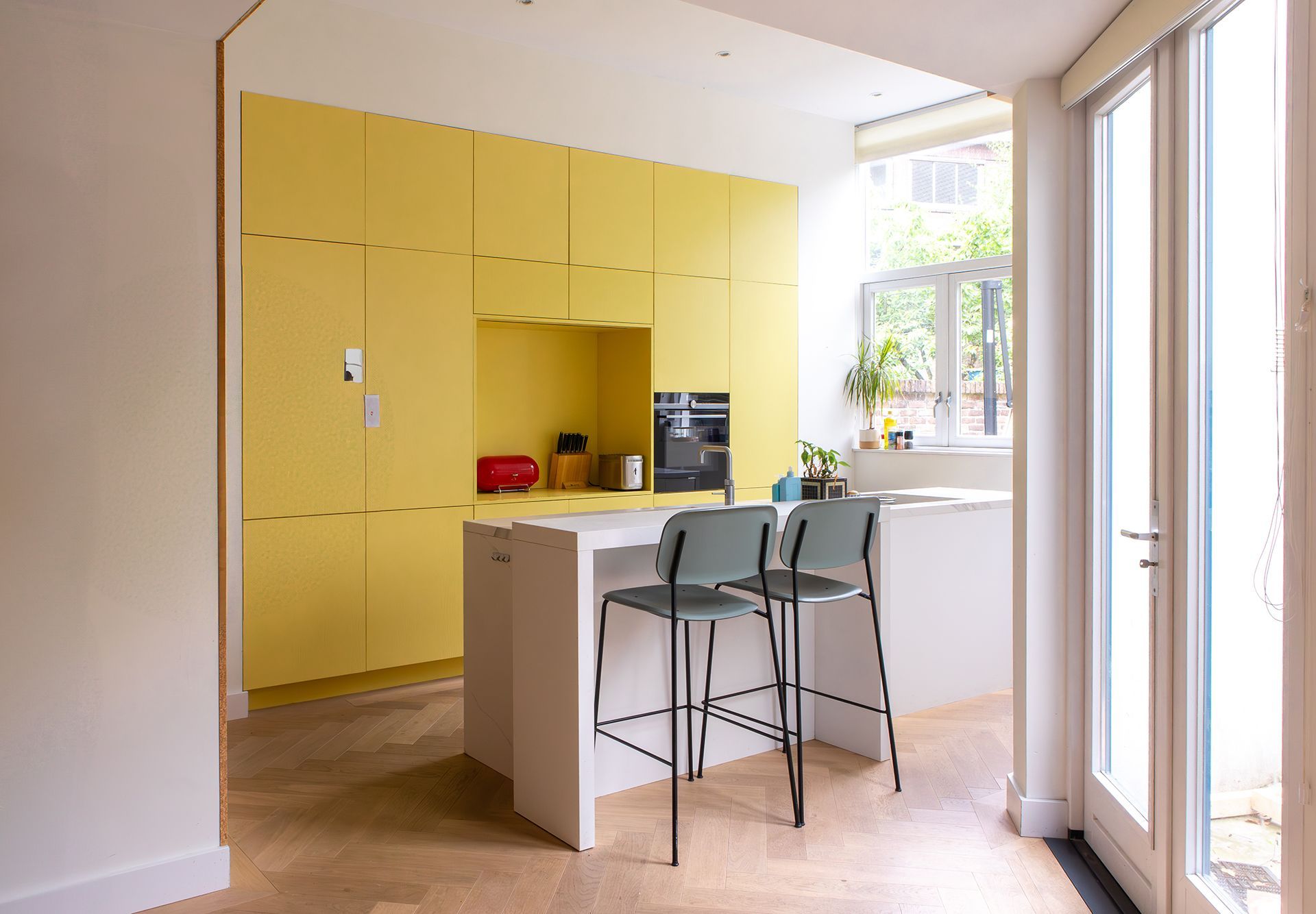 Gele keuken met wit kookeiland, turquoise barkrukken en een houten vloer met visgraatpatroon.