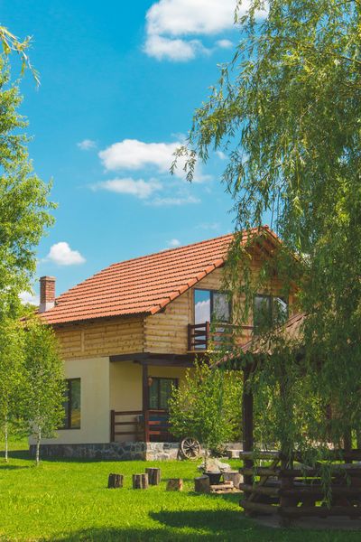 Two-story house with brown roof and light wood siding, nestled among green trees under a bright blue sky.