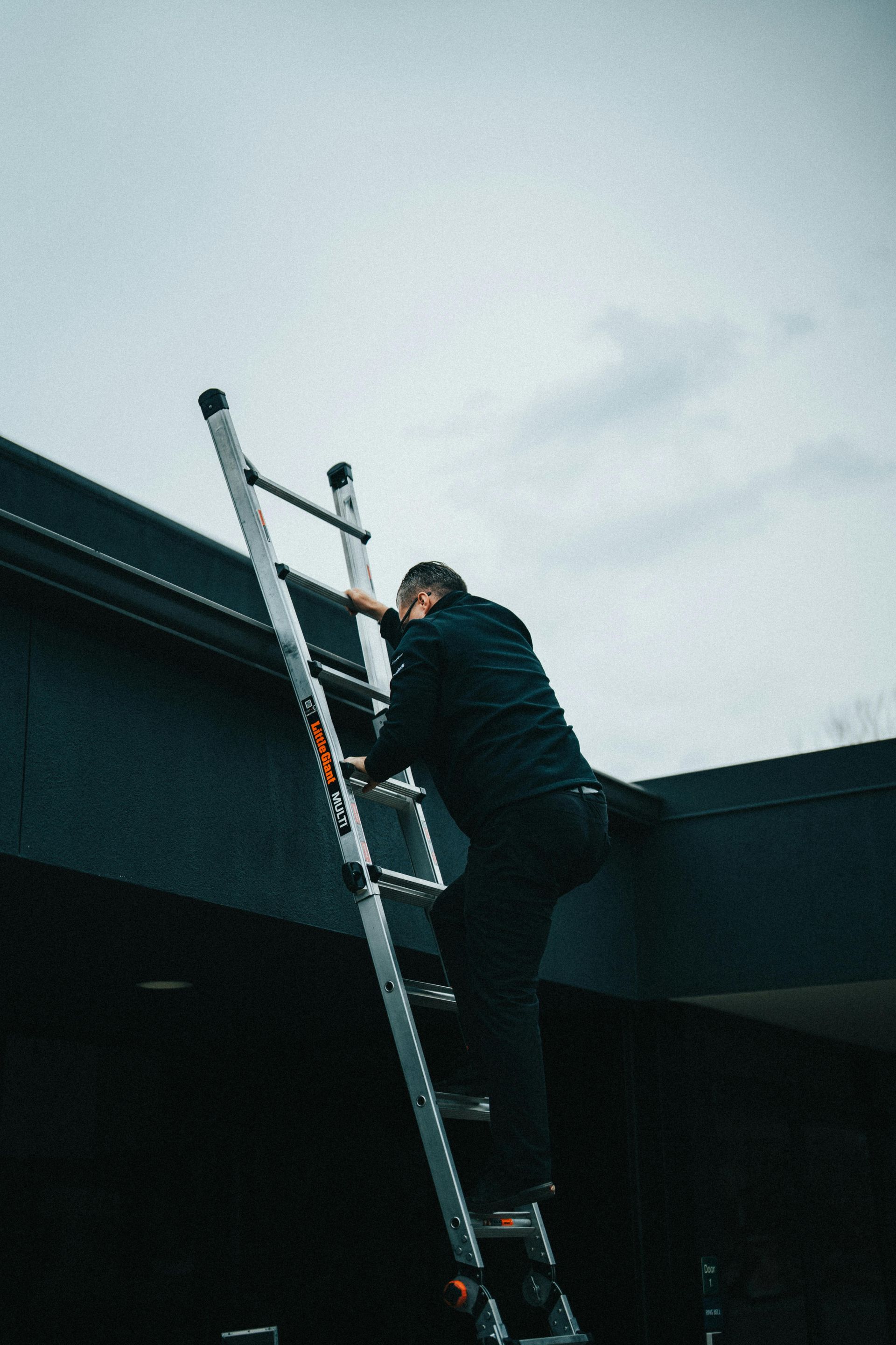 Man climbing a silver ladder to a dark-colored roof under a cloudy sky.