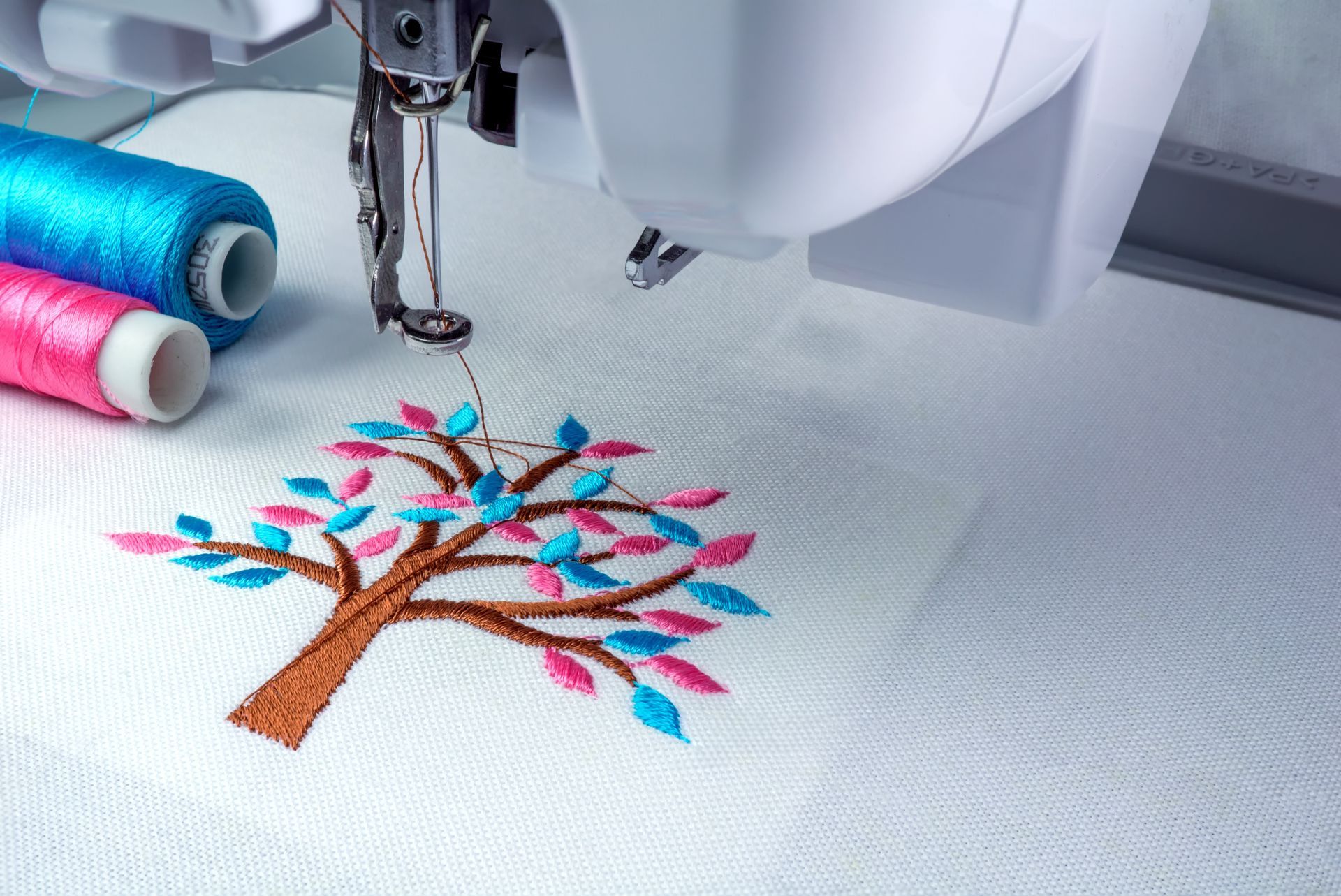 Embroidery machine stitching a pink and blue tree design on white fabric, with thread spools nearby