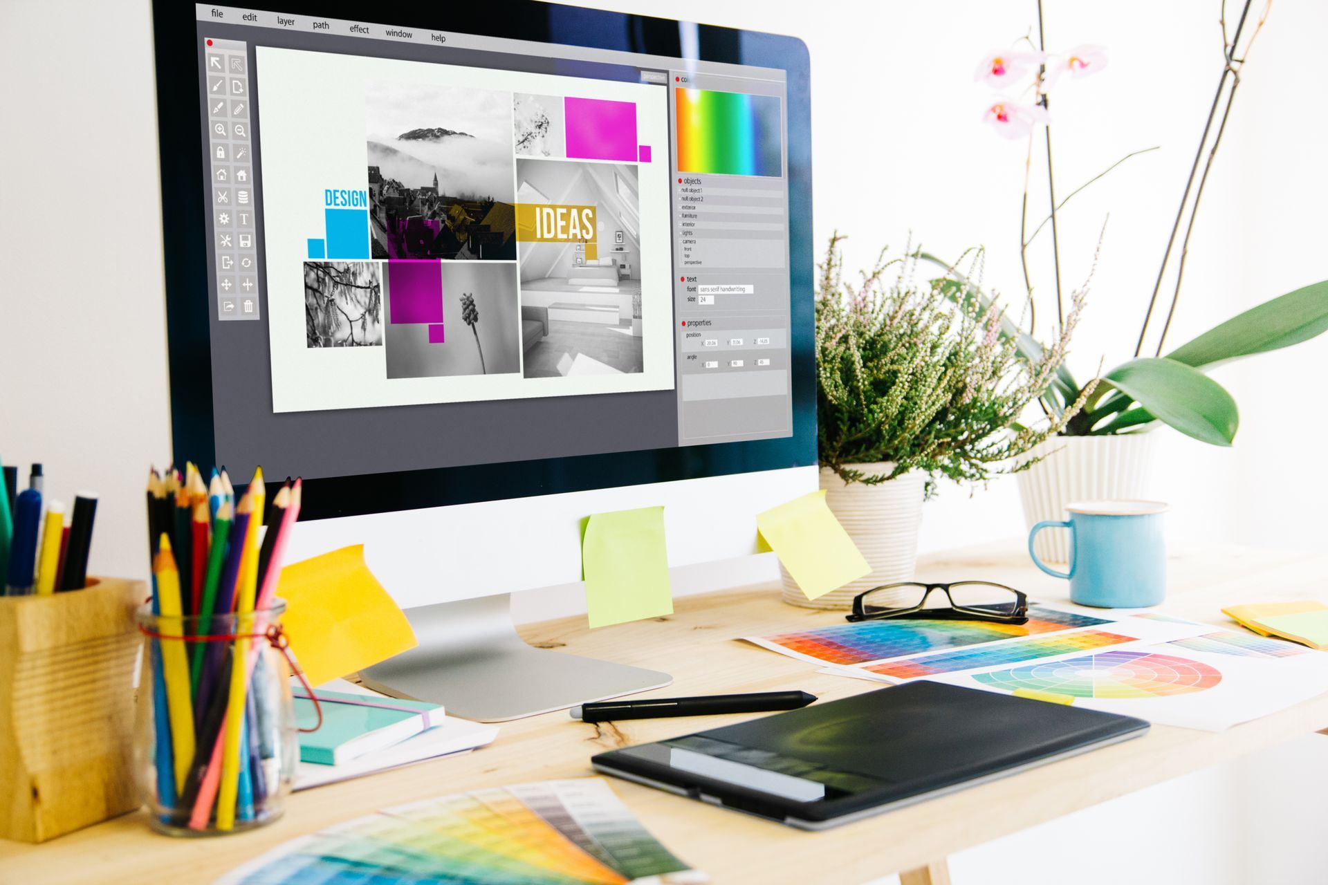 Creative workspace with iMac displaying a design layout, surrounded by colorful stationery and a potted plant