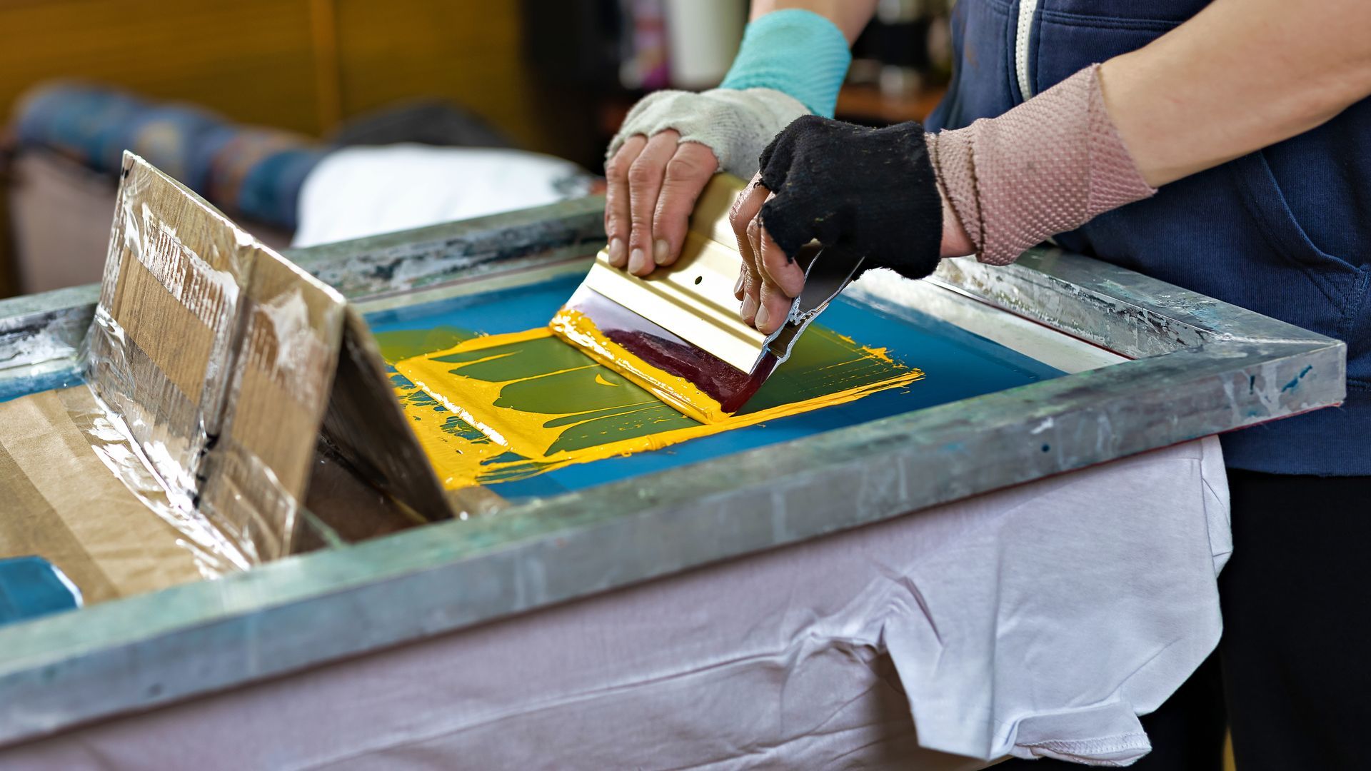Hands using a squeegee to screen print yellow and green paint on a fabric-covered frame