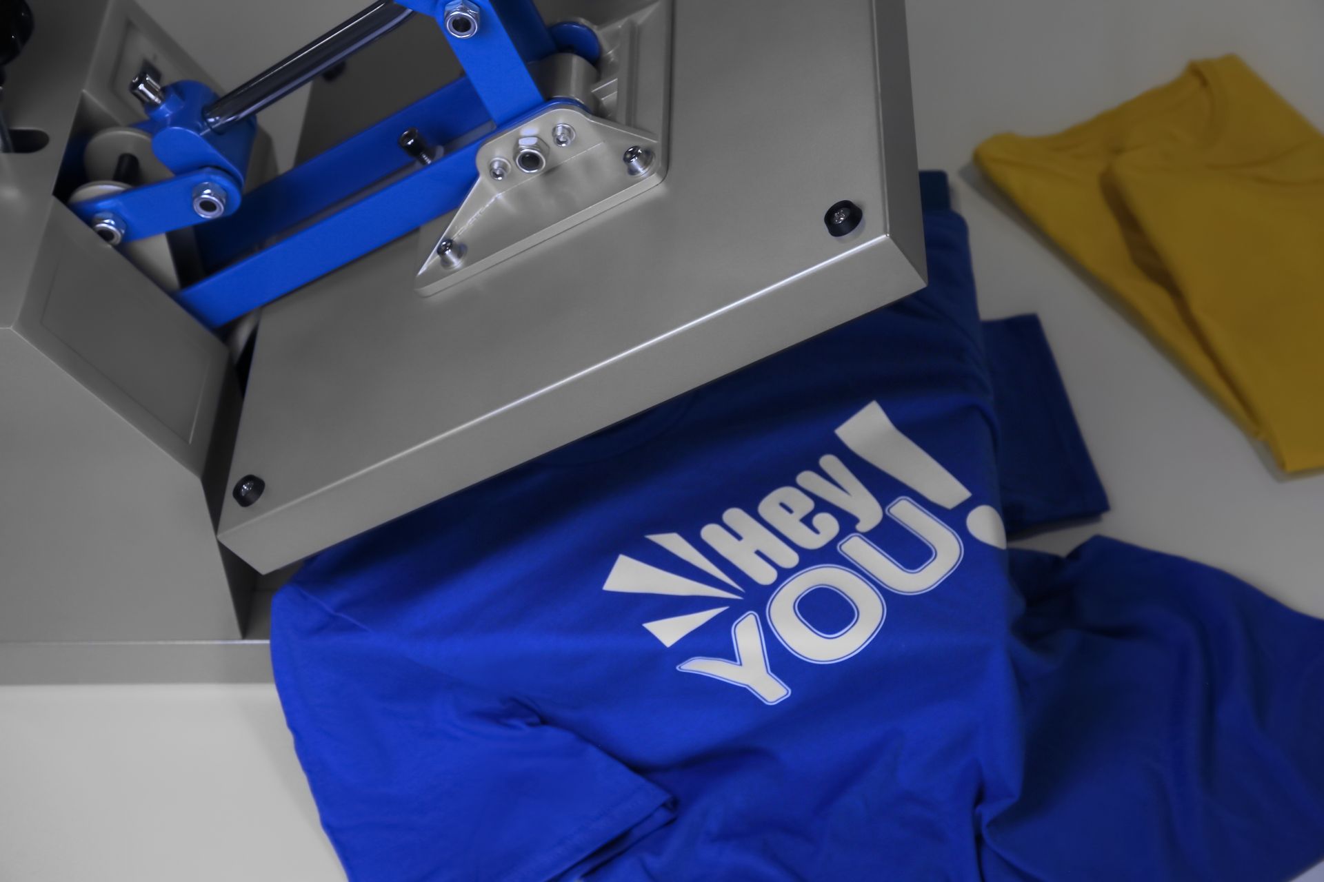 Blue T-shirt with “Thank You” text under a heat press, with a yellow shirt nearby.