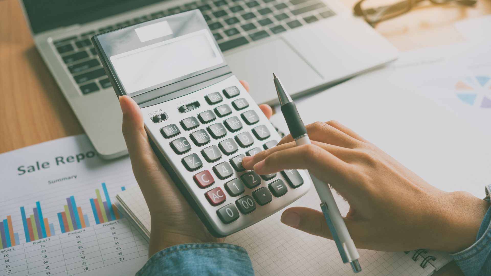 Person using a calculator, next to a laptop and charts, on a desk.