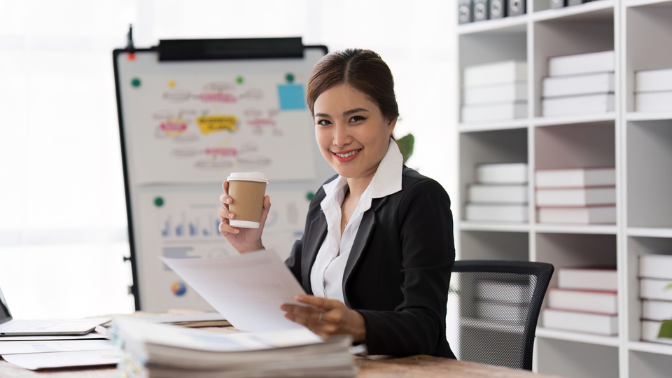 Businesswoman in black suit smiles, holding coffee and papers, at desk with whiteboard, shelves.