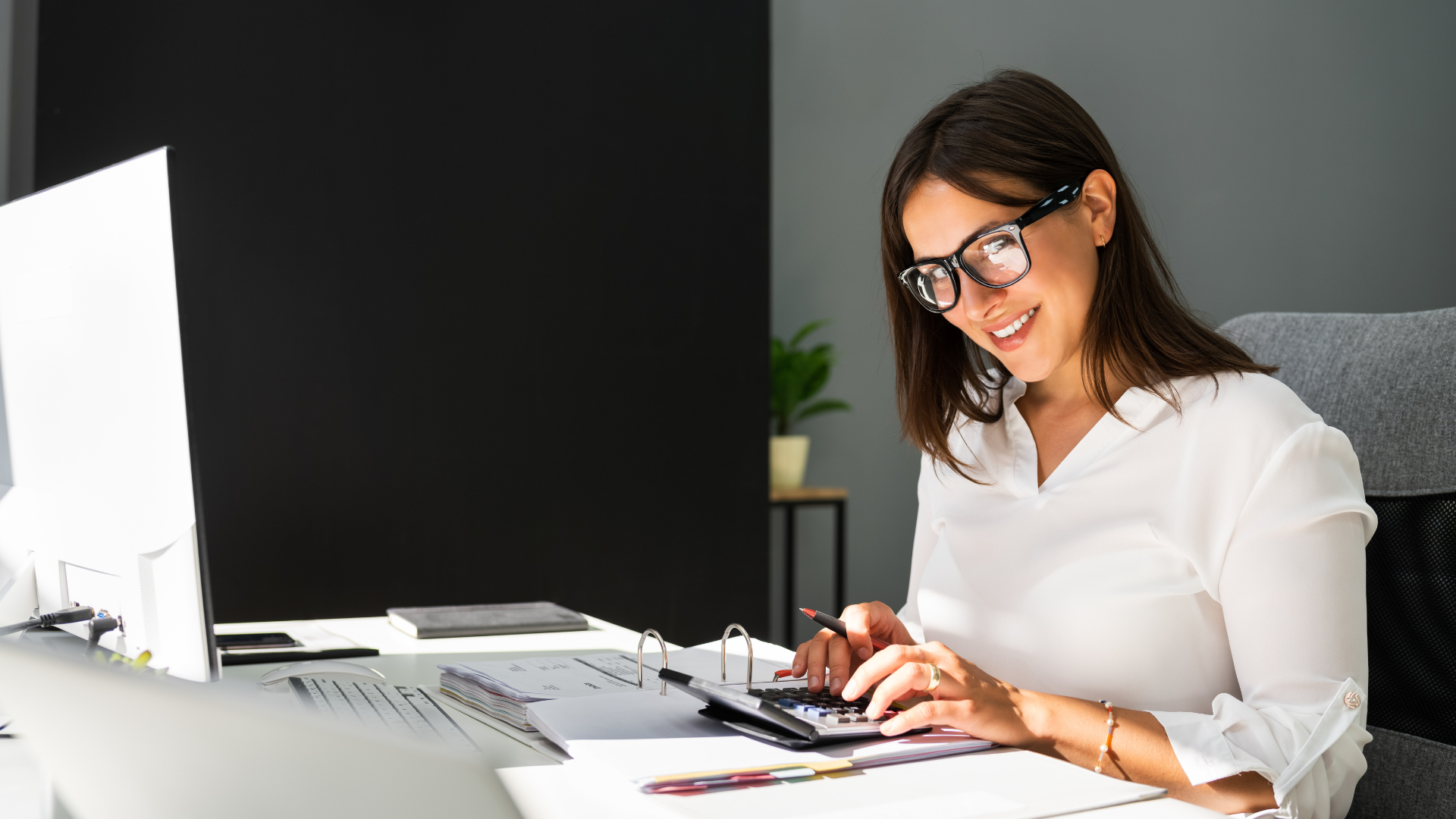 Woman wearing glasses smiling while calculating with a calculator at her desk.