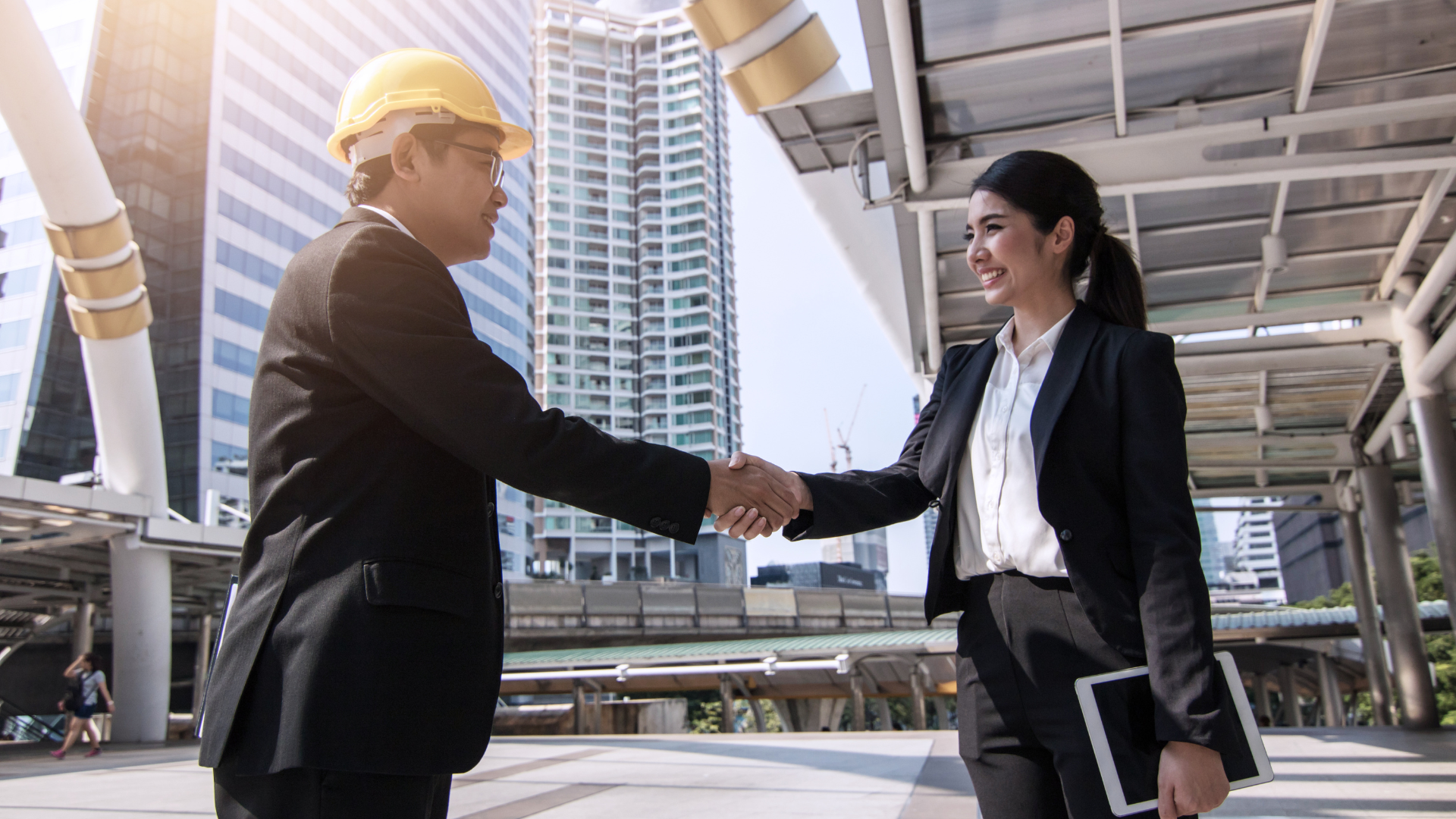 Man in hard hat and woman in suit shaking hands outdoors, city background.