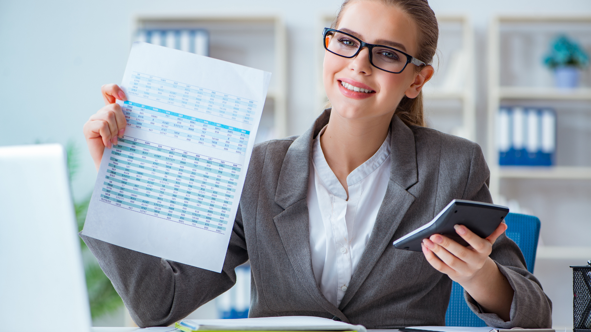 Businesswoman in glasses, holding a paper with charts and a calculator, smiling in an office setting.