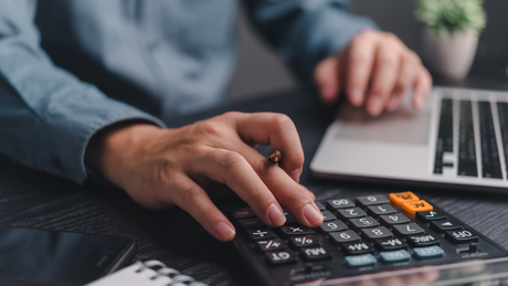 Person's hands using a calculator and laptop keyboard, calculating in an office setting.