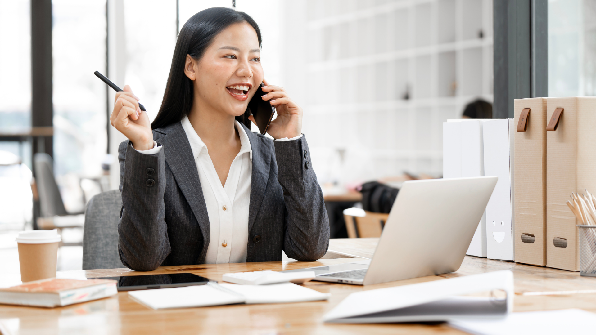 Woman in business attire smiles while talking on the phone in an office setting.