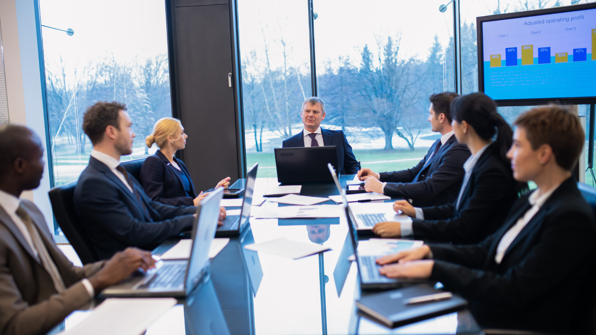 Business meeting in a modern office; a man leading, team working on laptops with a screen displaying a bar graph.