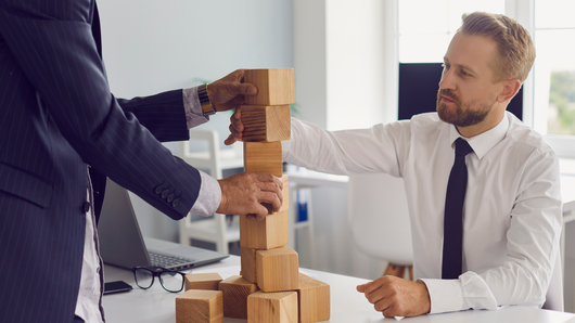 Two men in office playing a tower game with wooden blocks. One man's hand is on the tower, and the other man is