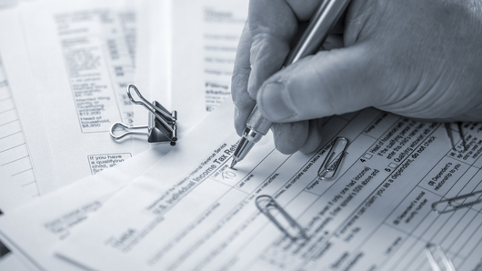 Hand writing on tax forms with a pen, alongside paperclips and a binder clip.