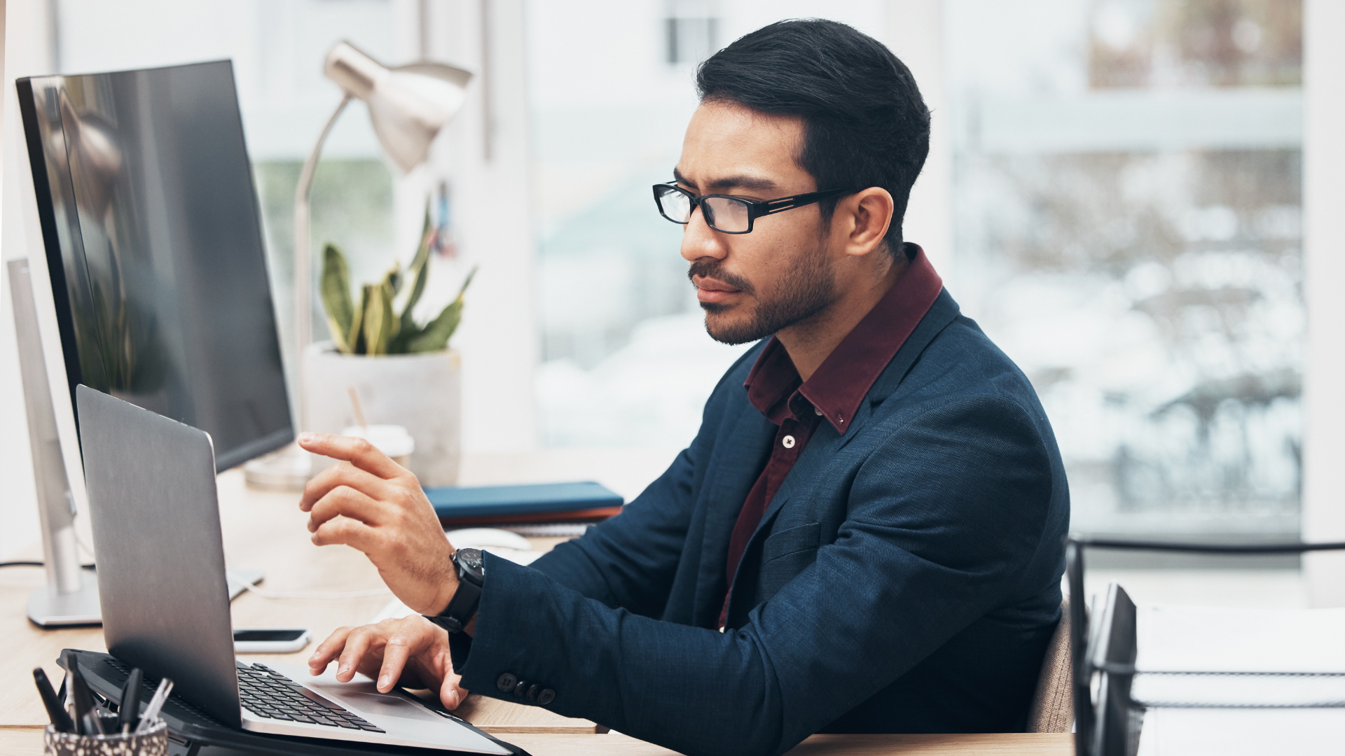 Man in glasses working on laptop and computer, in office setting.