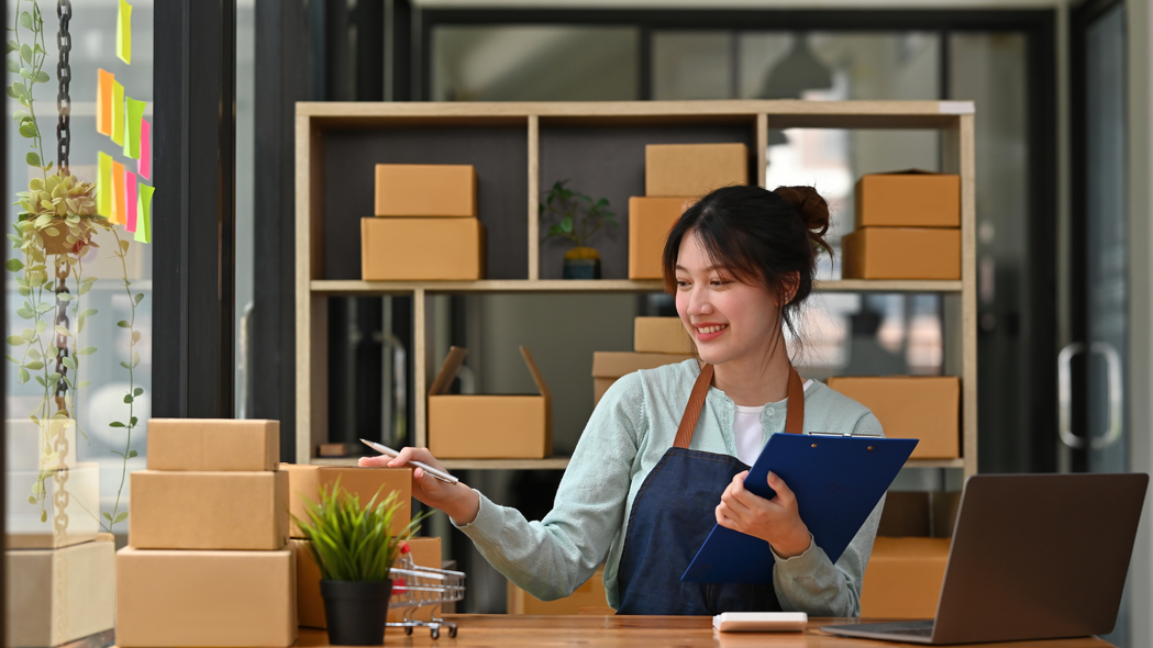 Woman in apron smiles, packing boxes in home office with shelves, laptop, and plants.