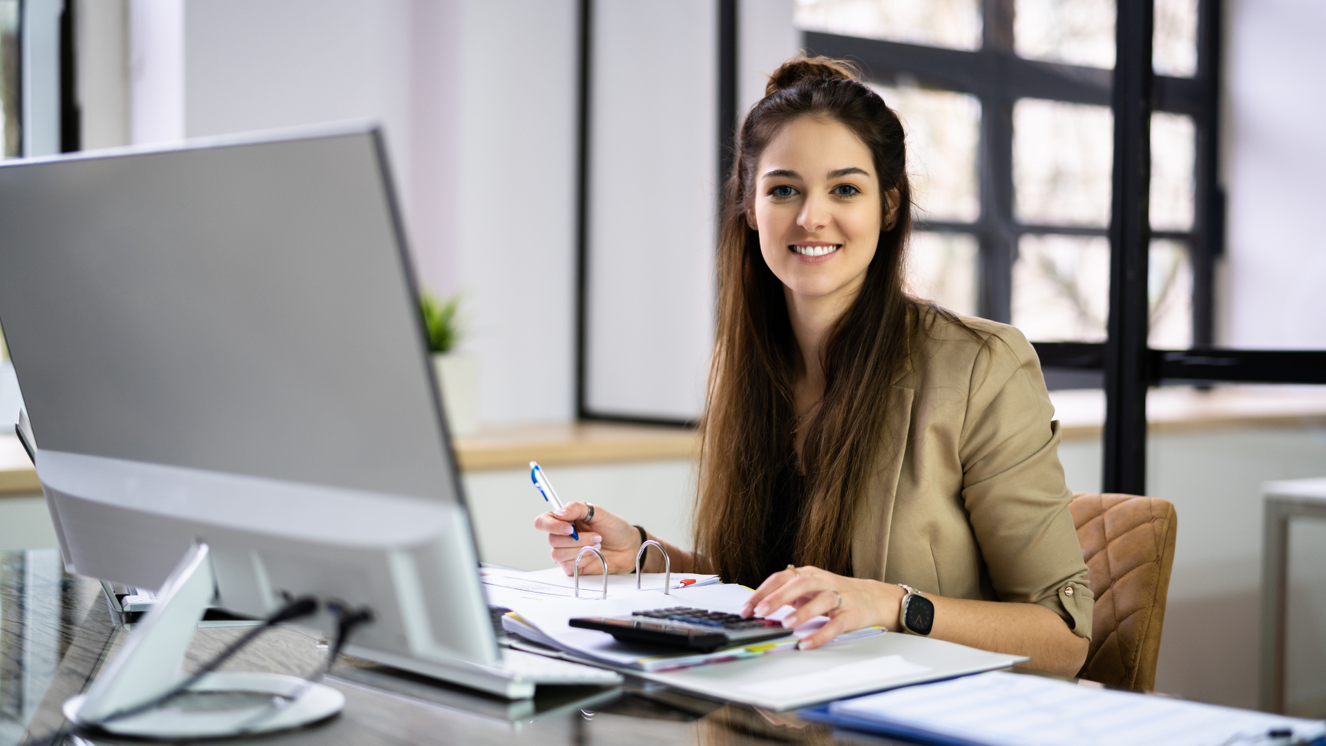 Woman in a blazer smiles while working on a computer in an office setting with a calculator and papers.