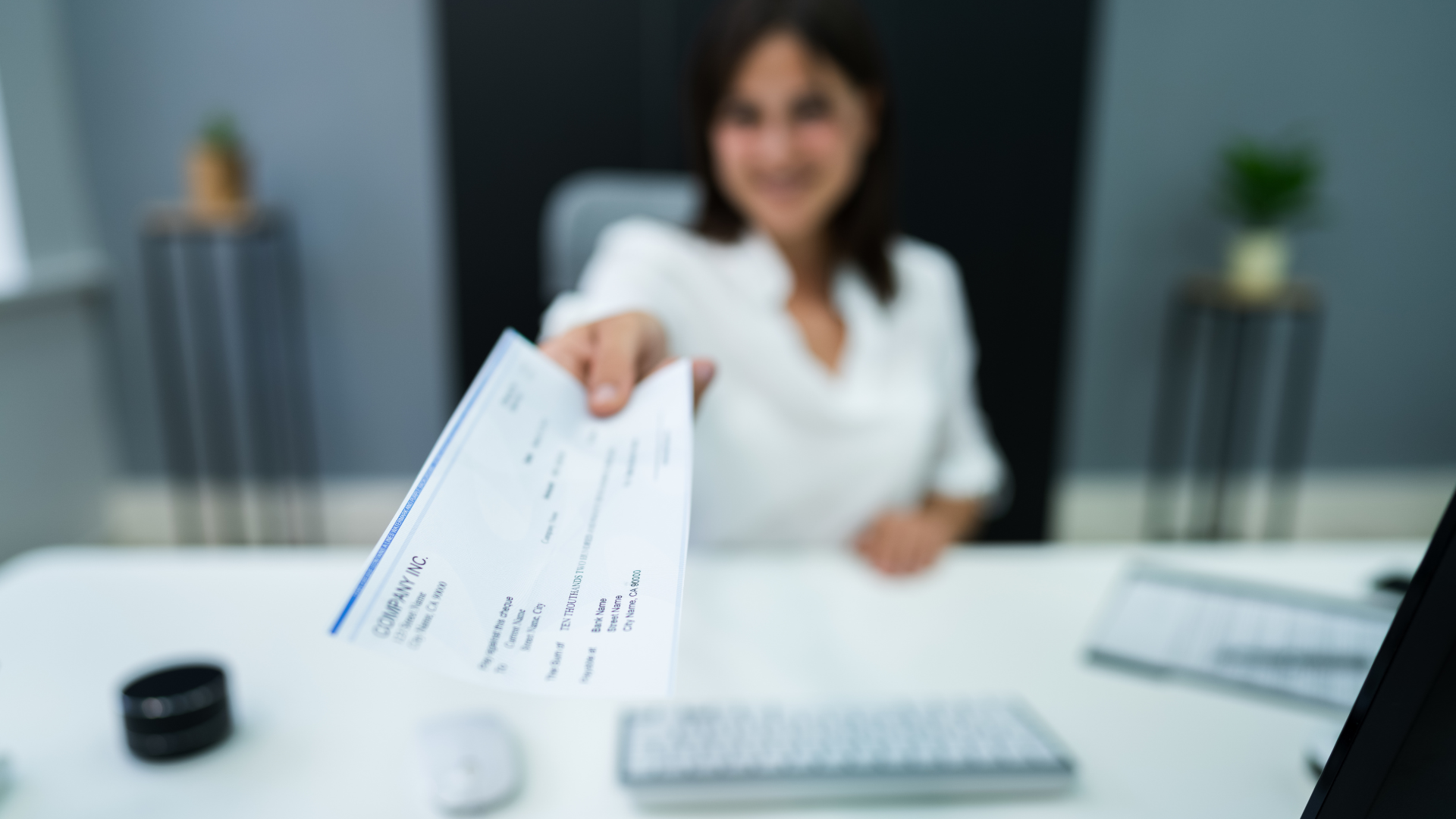 Woman in white shirt extends a check towards the viewer in an office setting, smiling.