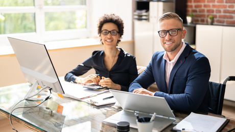 Two smiling people in a modern office, one in a wheelchair, working at a desk with a computer and laptop.
