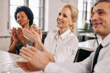 People clapping at a table. Smiling woman with blonde hair. Black woman clapping. Man smiling. Bright room.
