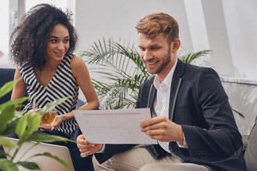 Woman and man in an office looking at papers, smiling, near a plant and window.