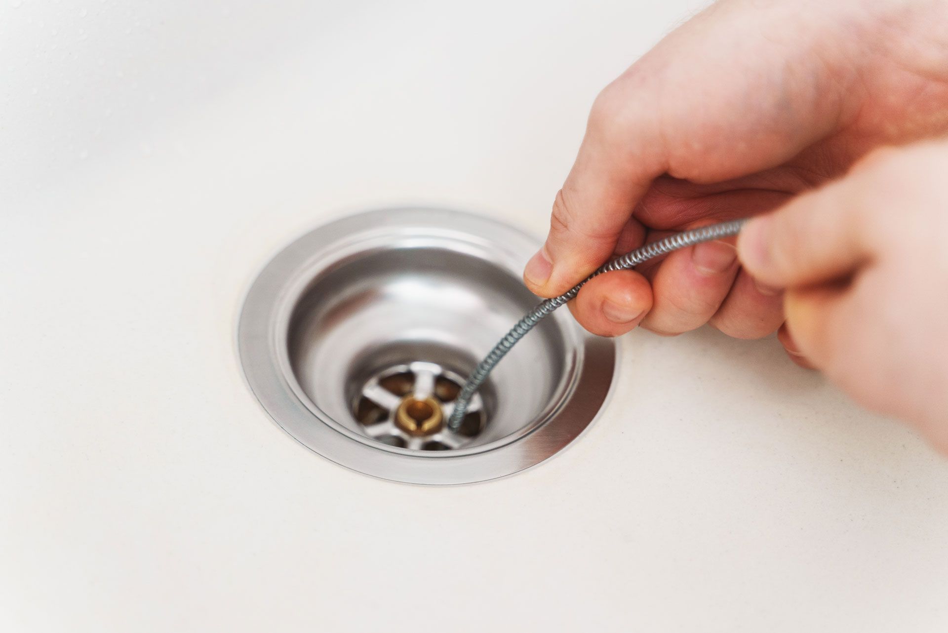 Hands using a drain snake to unclog a sink drain in a white sink. The drain is silver and round.