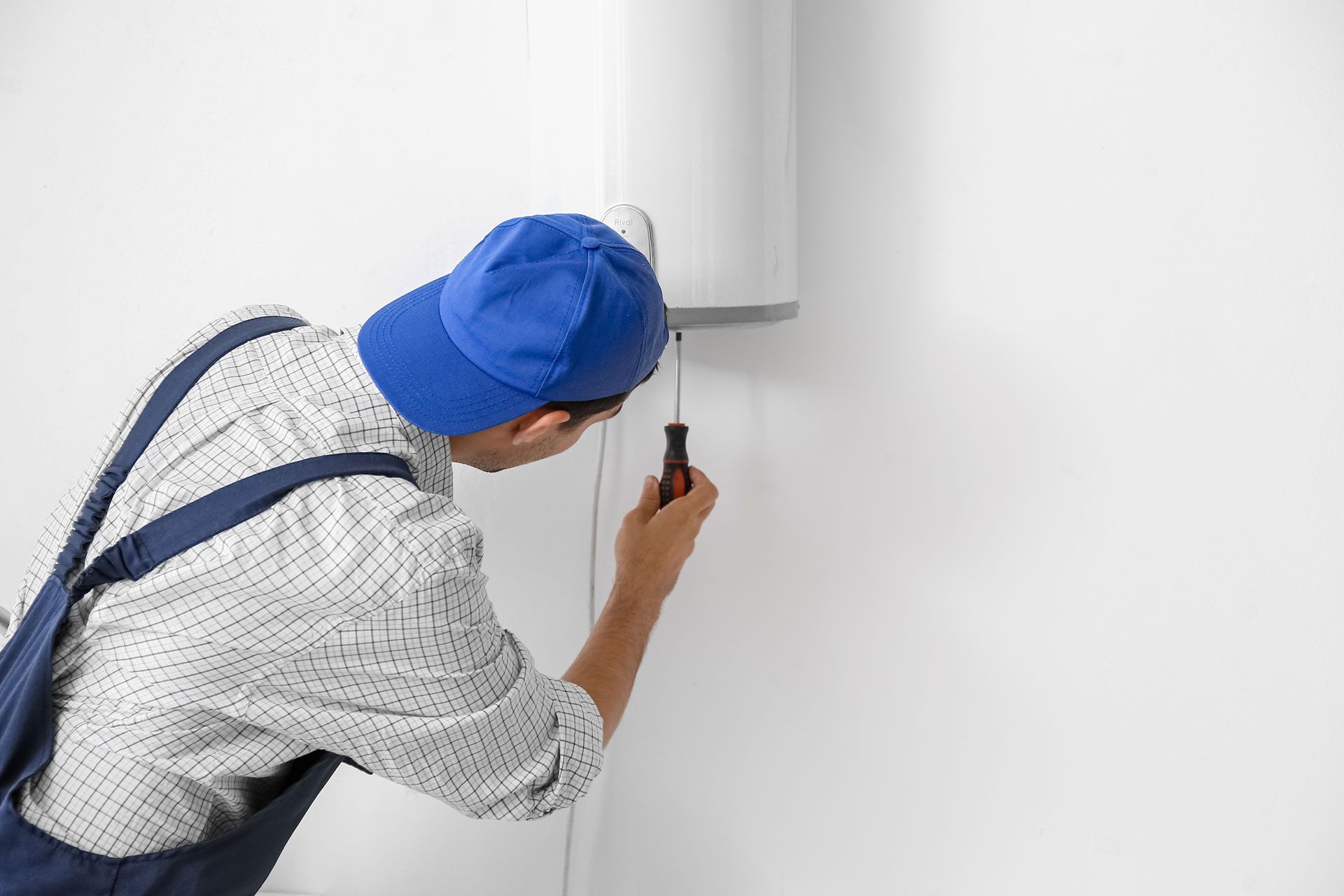 A person in a blue cap and overalls repairs a white water heater on a white wall, using a screwdriver.