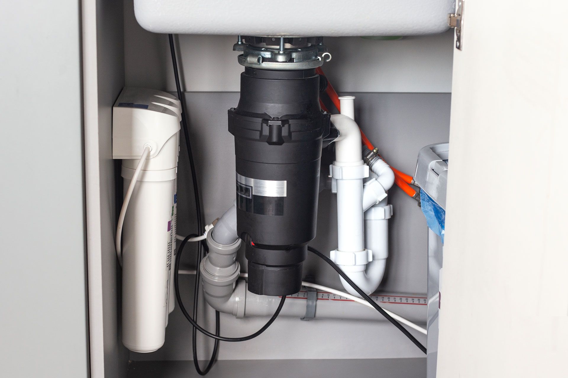 Under-sink view with garbage disposal, plumbing, and water filter in a gray cabinet.