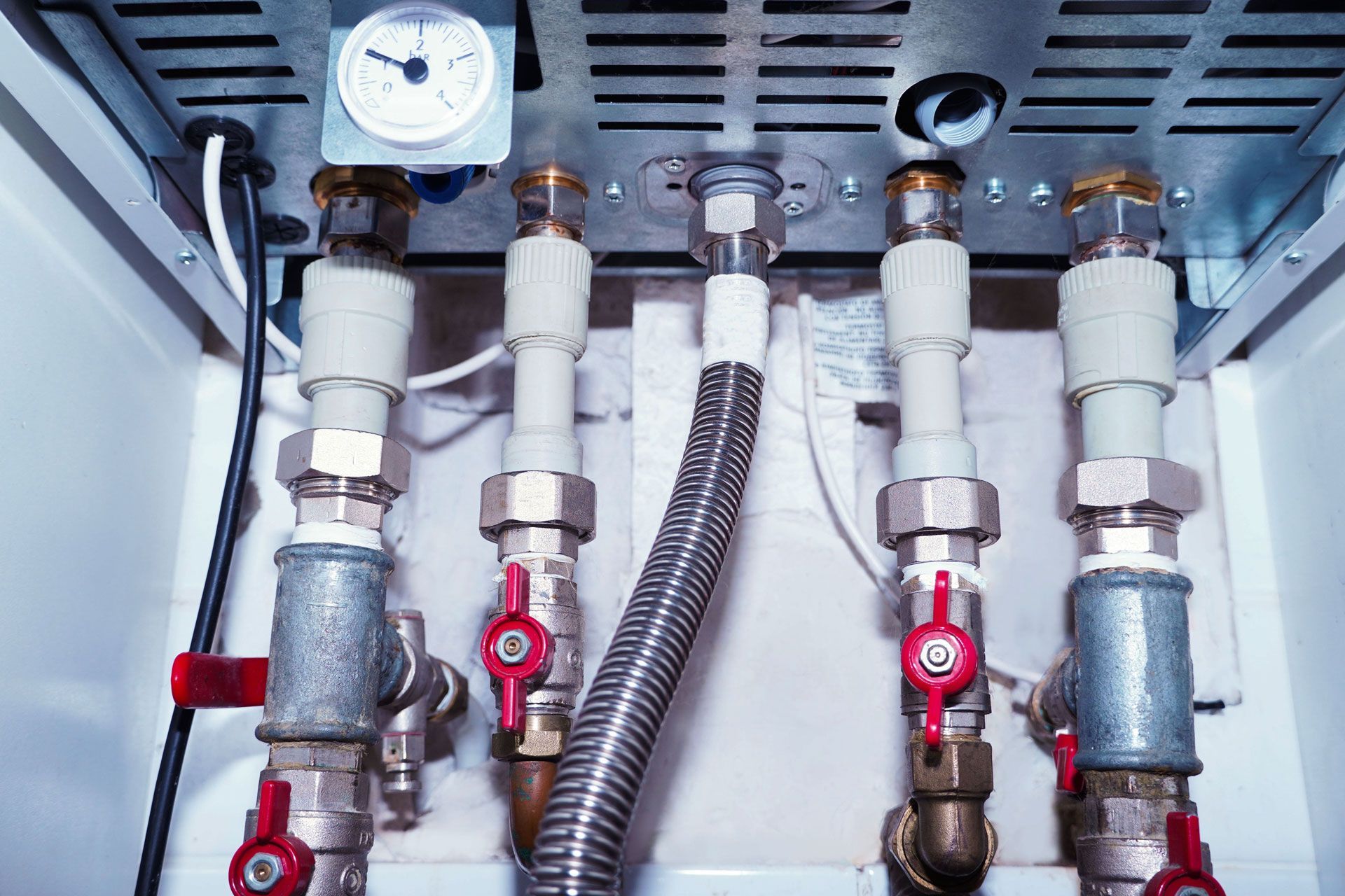 Close-up of a boiler's plumbing with pipes, valves, a gauge, and flexible tubing, inside a white enclosure.