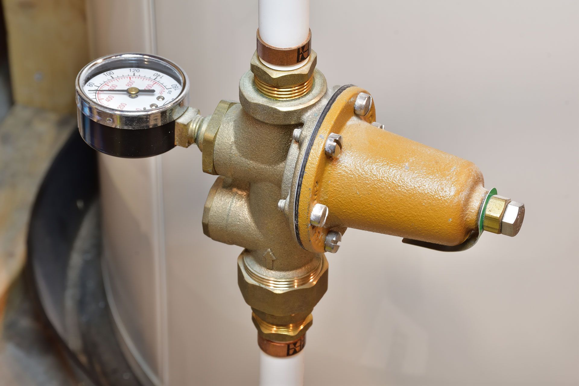 A pressure regulator with a gauge attached to white pipes. The regulator is bronze, and the gauge reads the water pressure.
