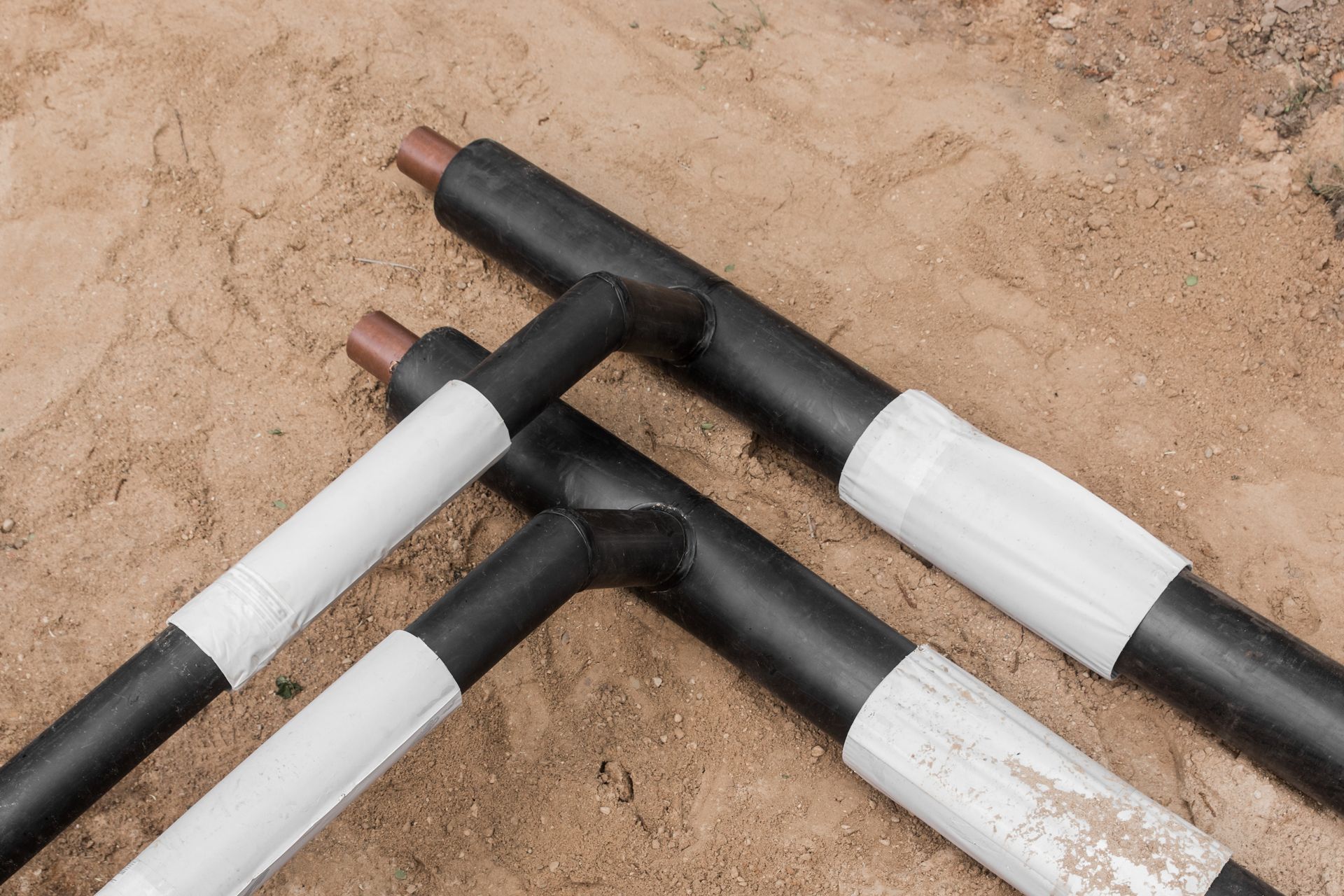 Black insulated pipes with brown ends and white tape on sand.
