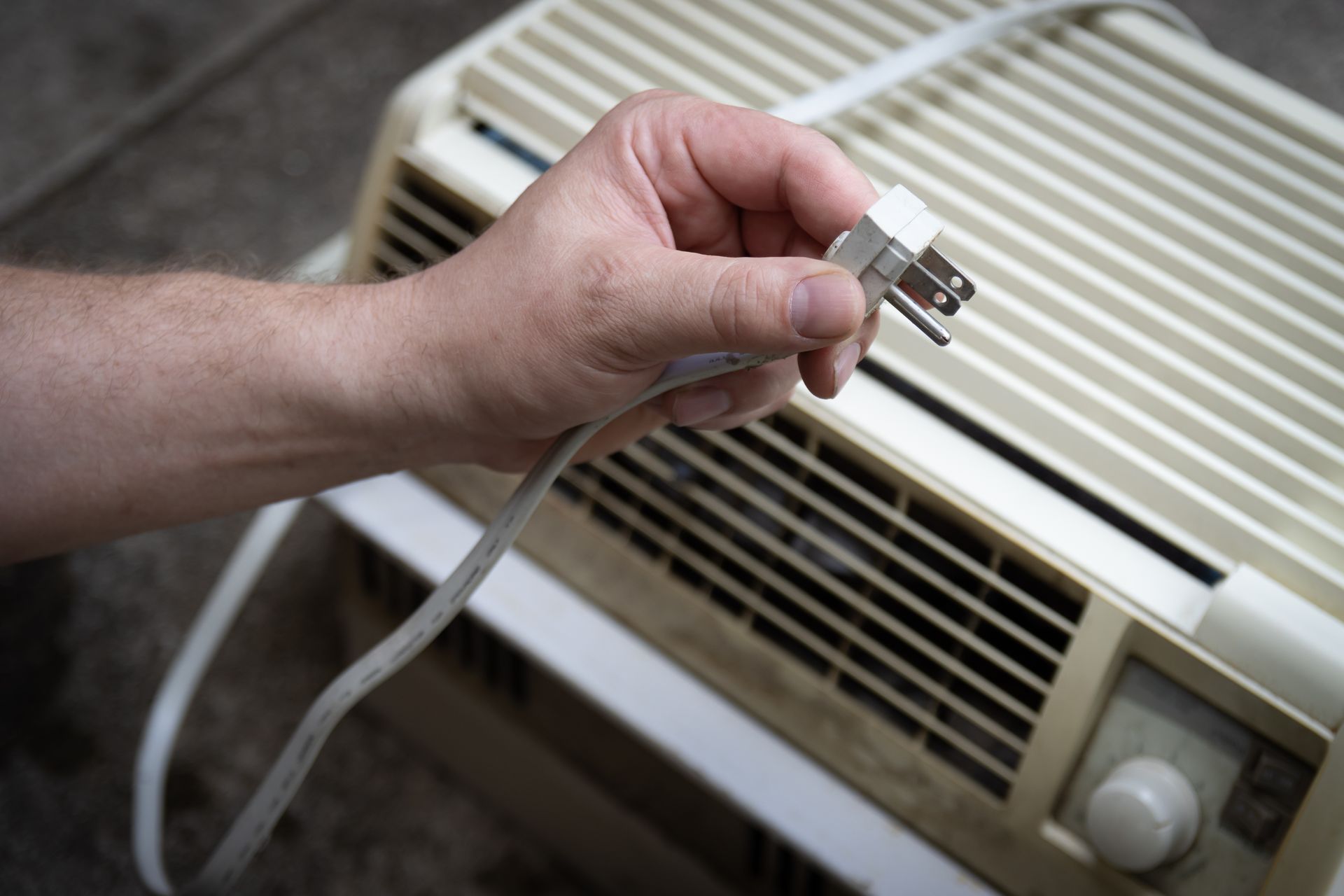 Hand plugging a cord into a window air conditioner. The unit is beige.