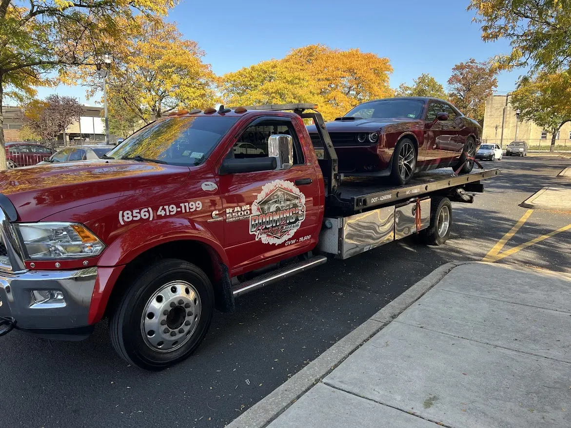 Red tow truck carrying a dark car on a sunny day near trees with fall foliage.