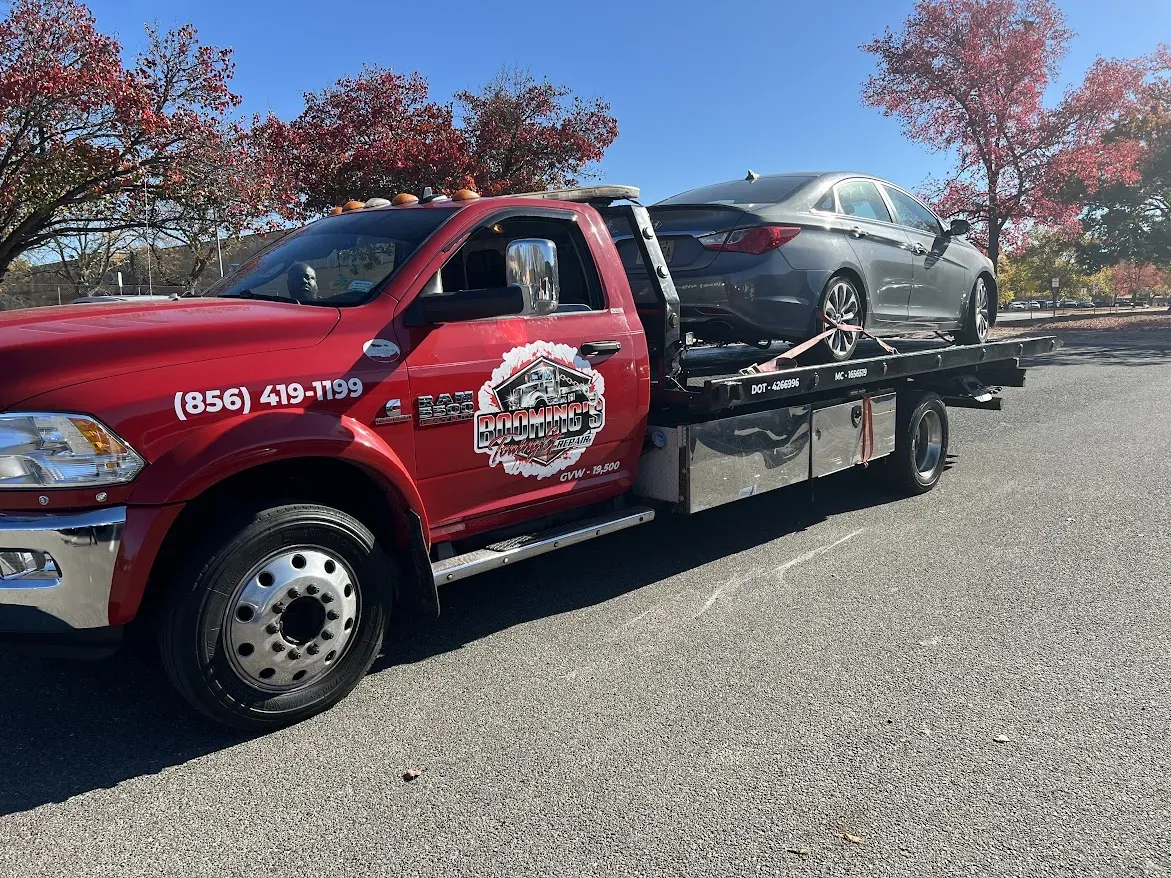 Red tow truck carrying a gray car on a sunny day; trees with red leaves in background.