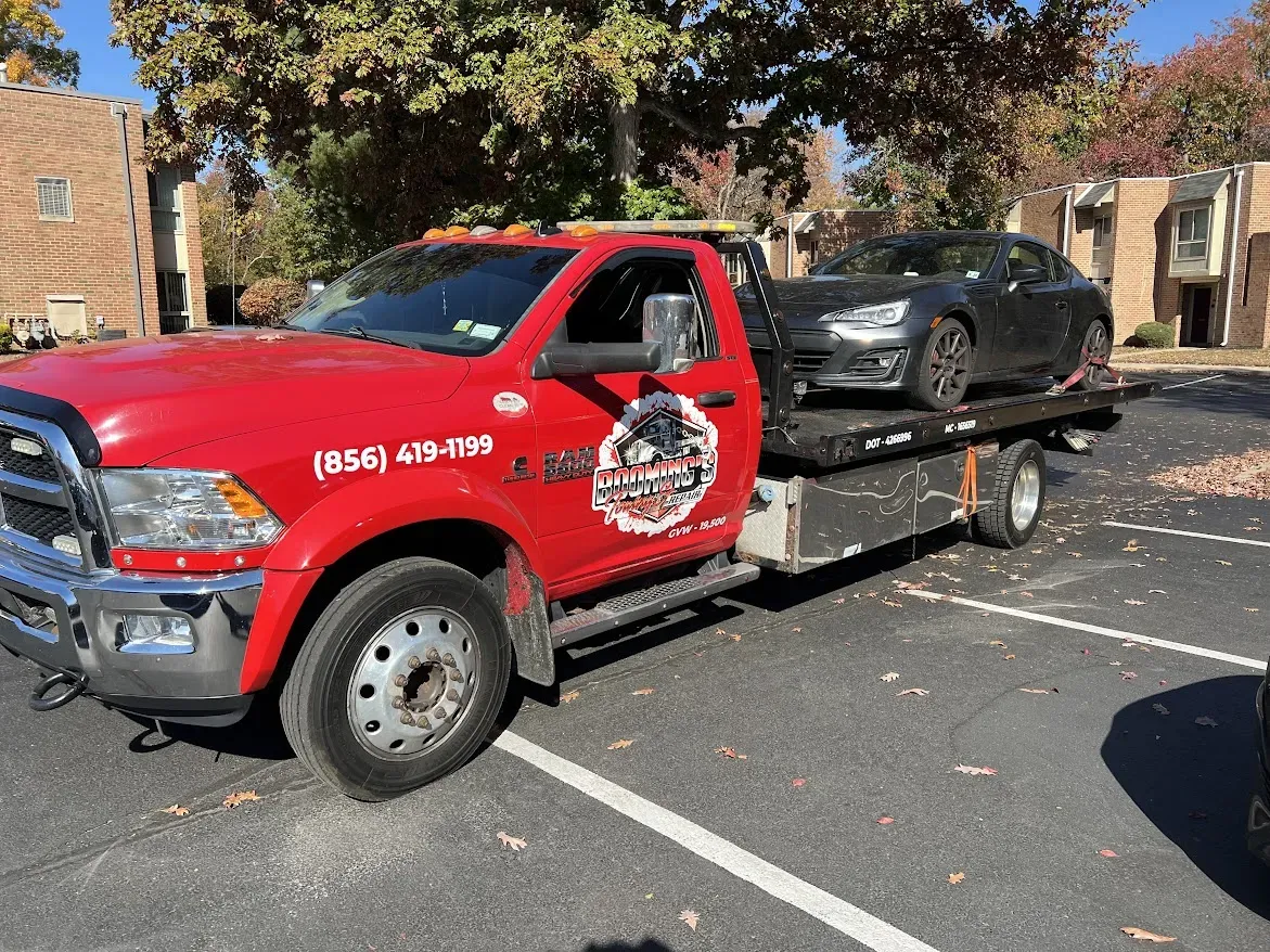 Red tow truck towing a black car in a parking lot, autumn trees in background.