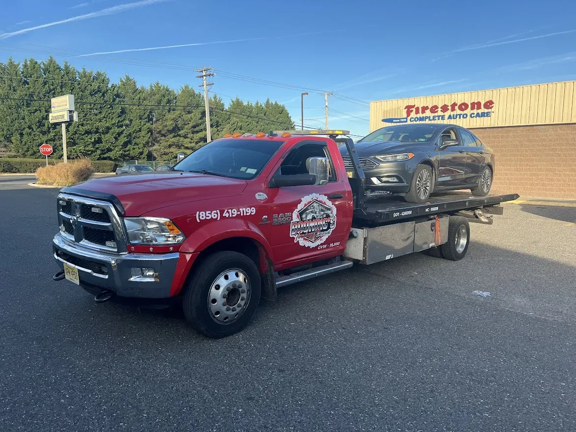 Red tow truck hauling a gray car in front of a Firestone store.