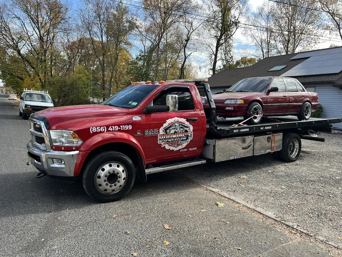 Red tow truck carrying a maroon sedan on a flatbed. Sunny outdoor setting.