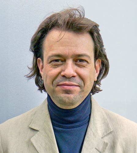 Ein Mann mit langen Haaren trägt eine hellbraune Jacke und einen blauen Rollkragenpullover.