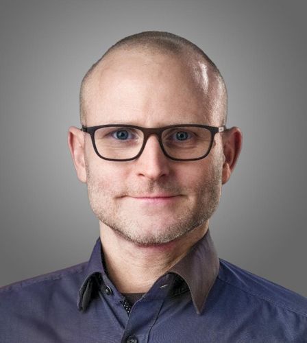 Ein kahlköpfiger Mann mit Brille und blauem Hemd lächelt.