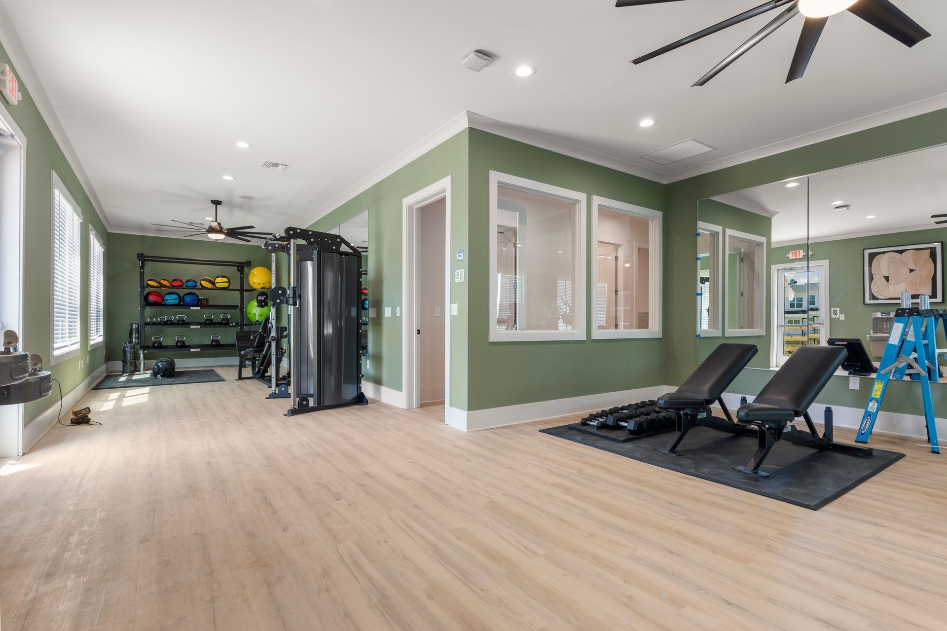 Bright home gym with light wood floors, green walls, exercise equipment, and two reclining benches under ceiling fans