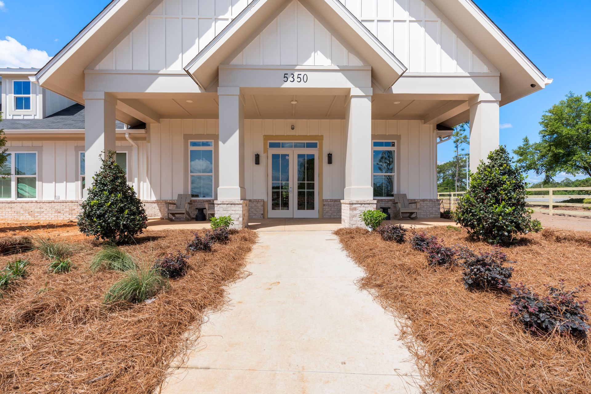 Front entrance of a beige house with a white porch and landscaped mulch beds along a concrete walkway