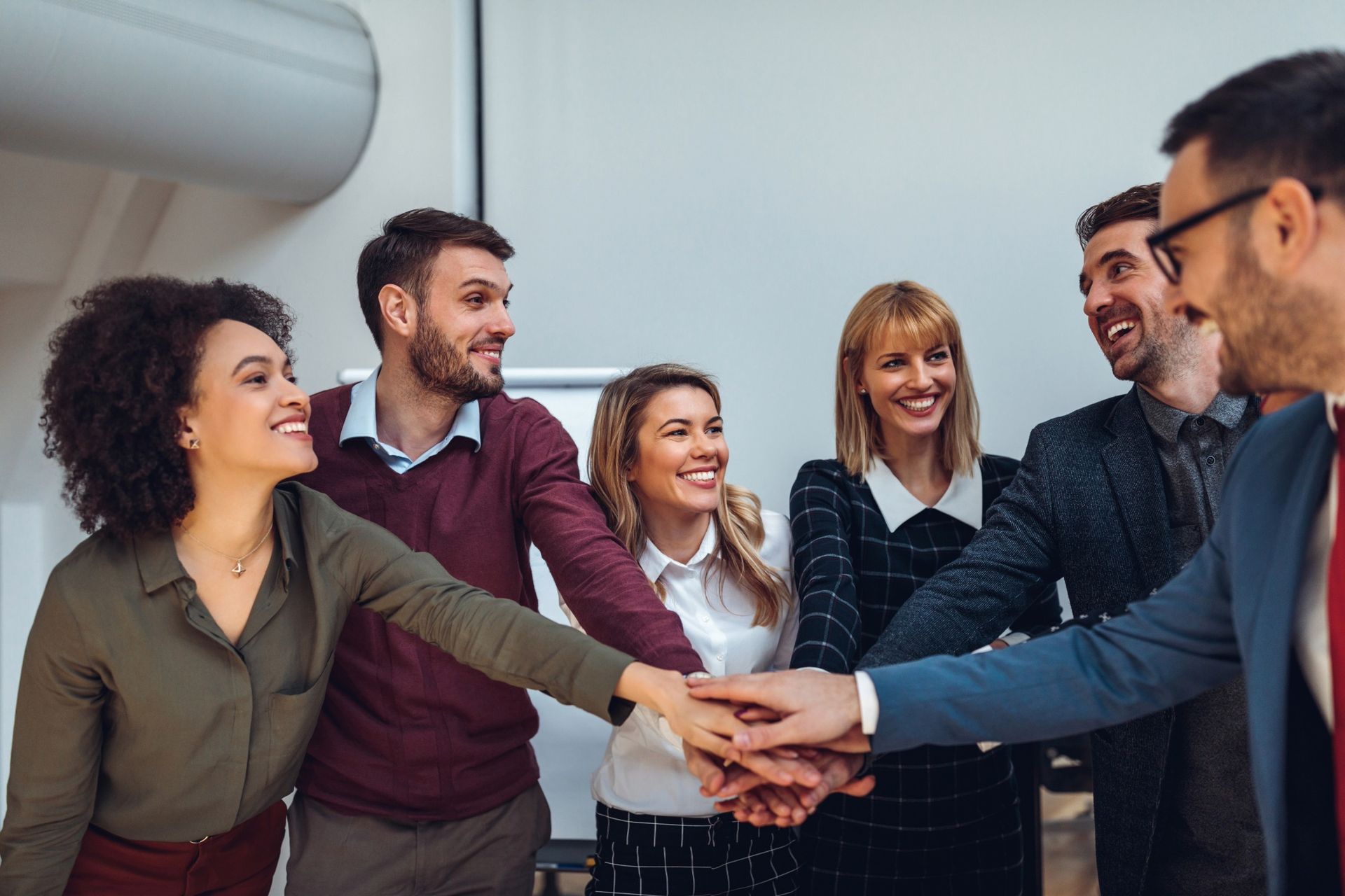 A group of professionals in an office setting smiling as they stack their hands together in a sign of teamwork.