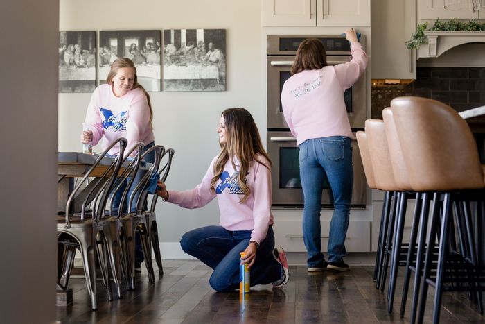 Three people in matching pink shirts clean a kitchen, with one wiping chairs, one scrubbing a wall, and one at an oven.
