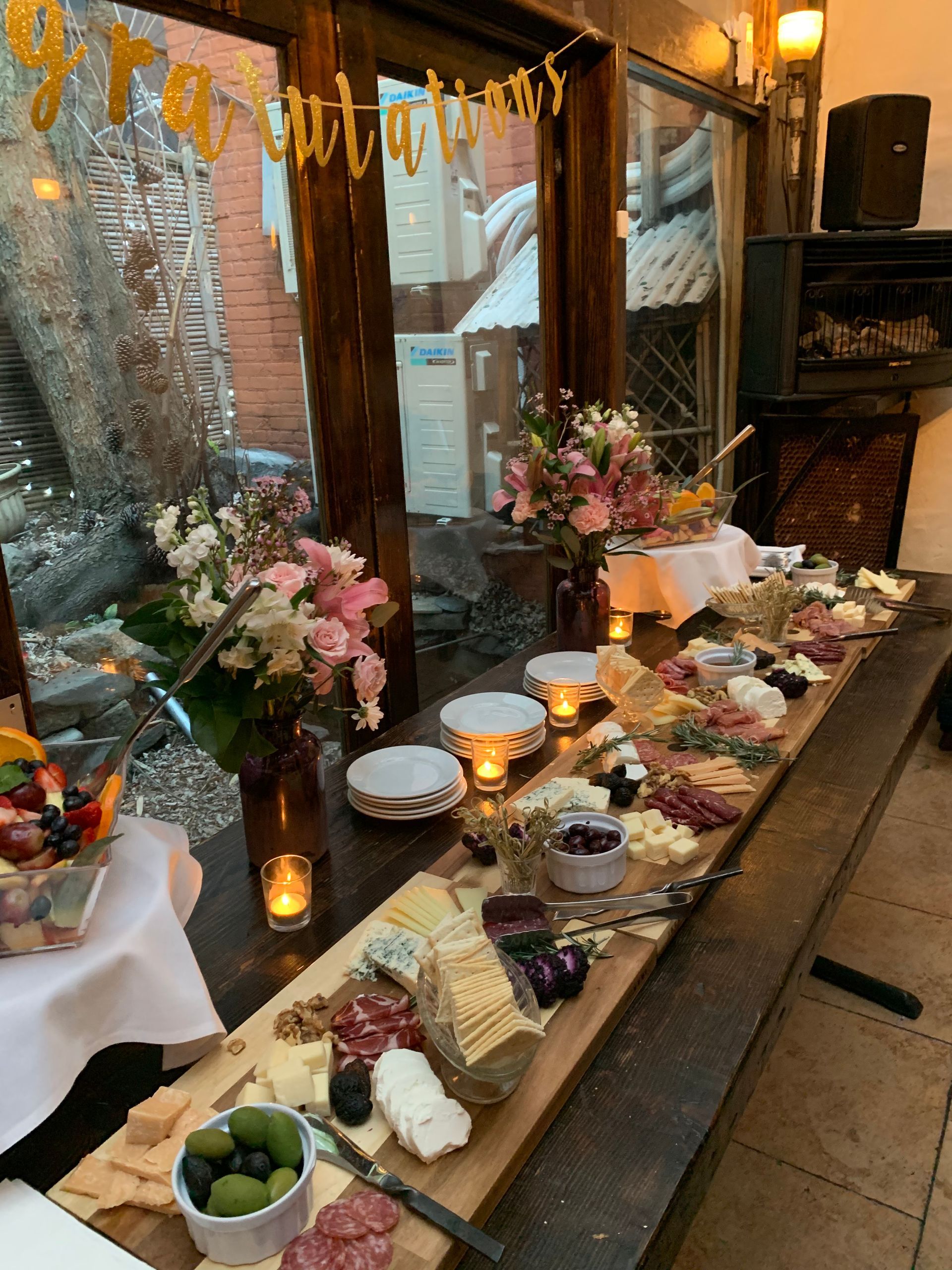 A long charcuterie table featuring meats, cheeses, crackers, olives, and floral arrangements, with a congratulations banner.