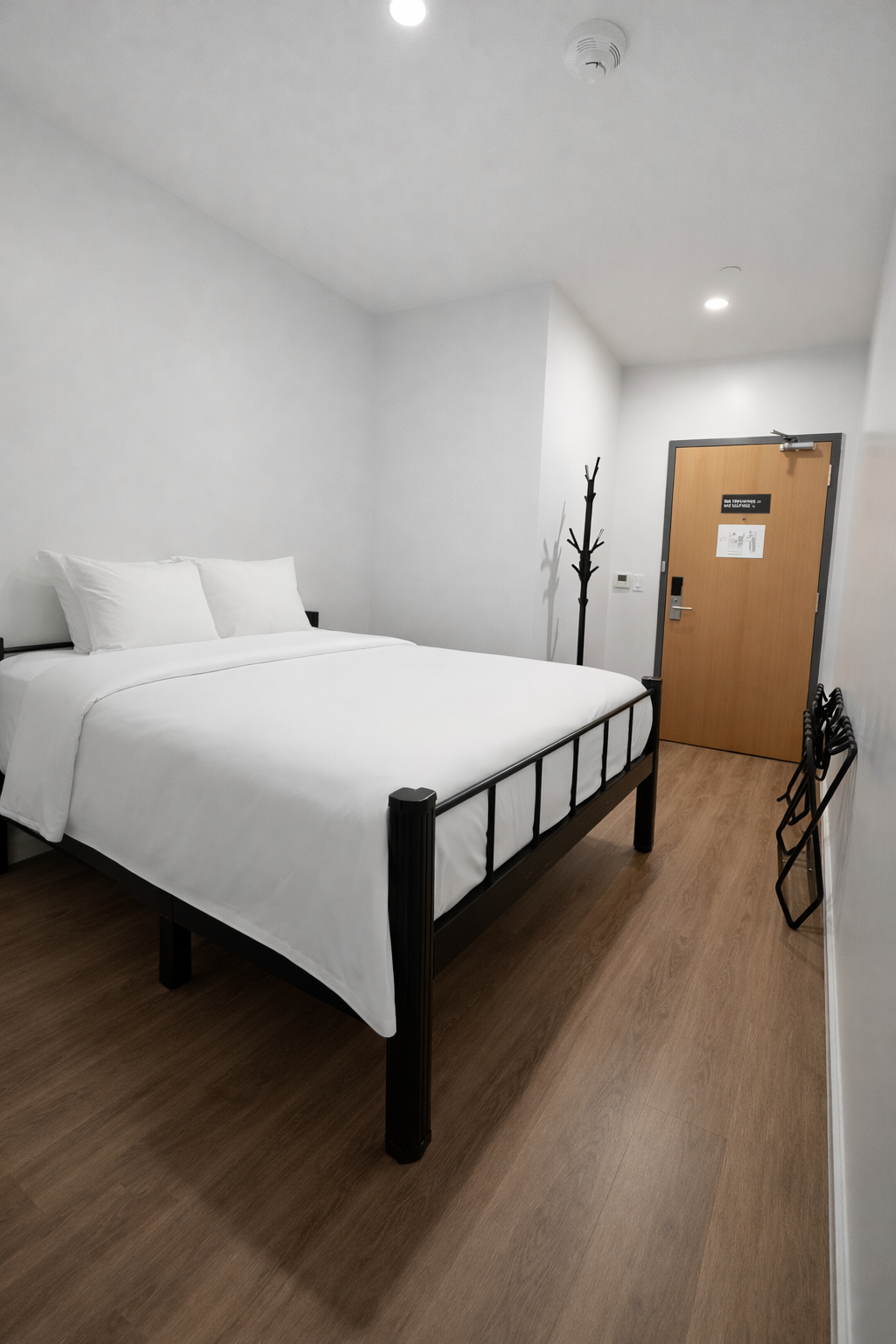 A hotel room with a queen bed featuring a black metal frame, white linens, and light wood flooring leading to a door.