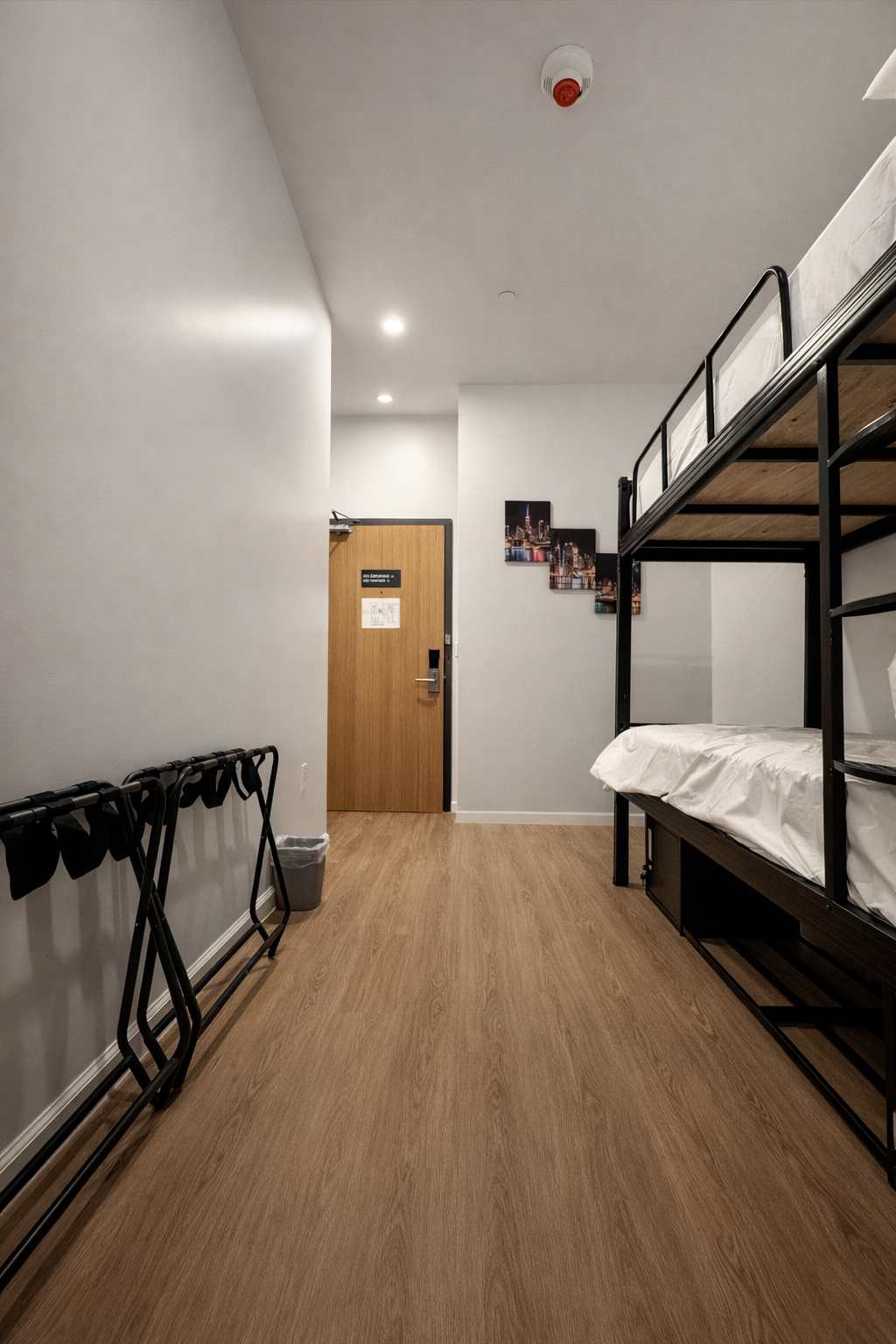 A hallway in a hostel room with a black metal bunk bed on the right, light wood floors, and a door at the end.