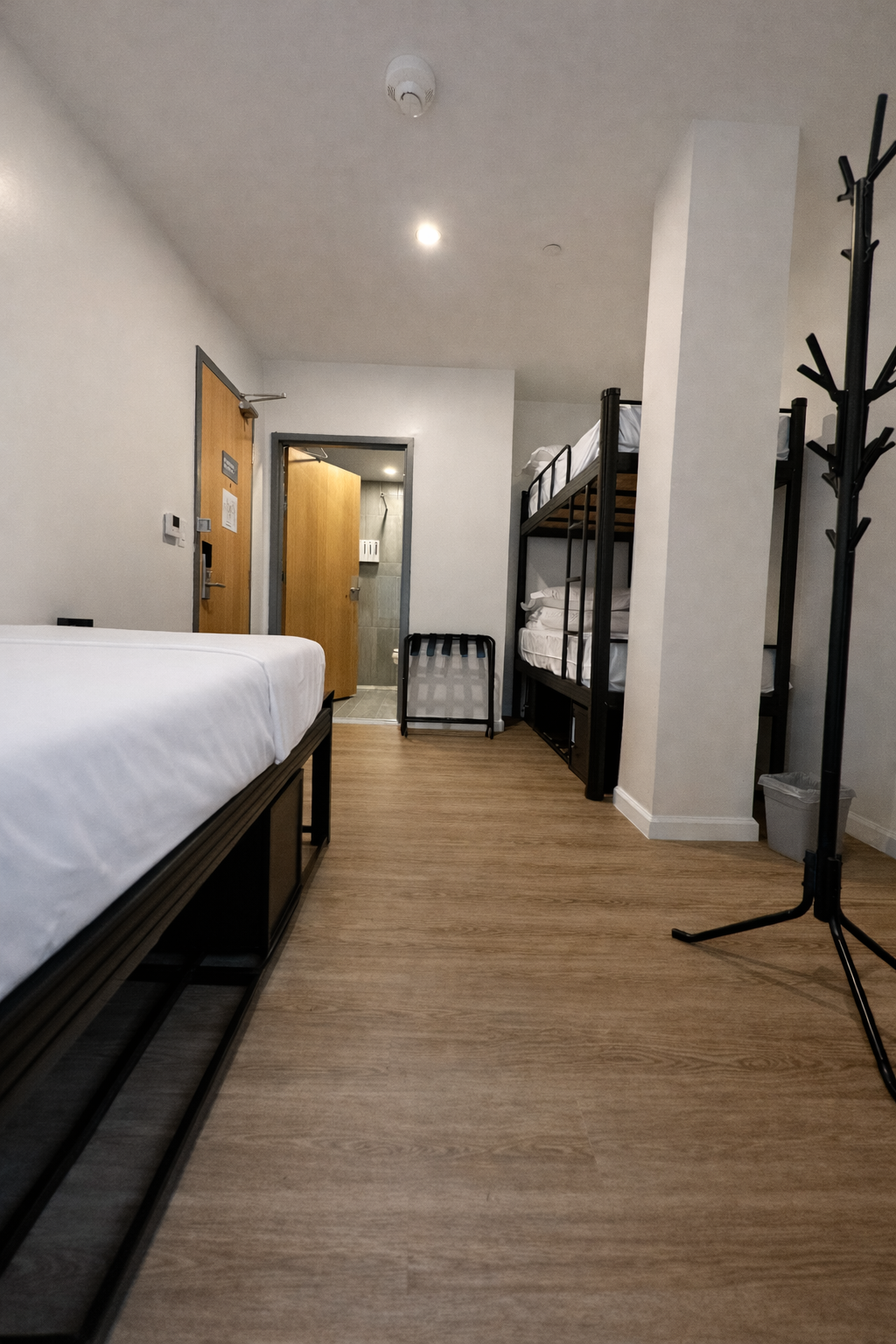 A hotel room with a foreground bed, a bunk bed in the background, a coat rack, and a doorway leading to a bathroom.