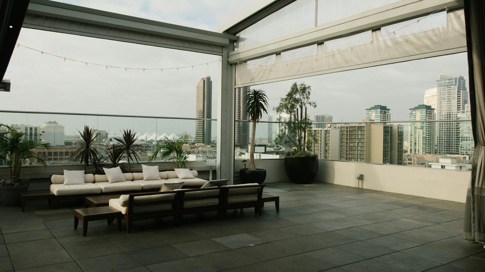 Rooftop patio with city view: sofas, plants, and a retractable roof. Overcast sky.