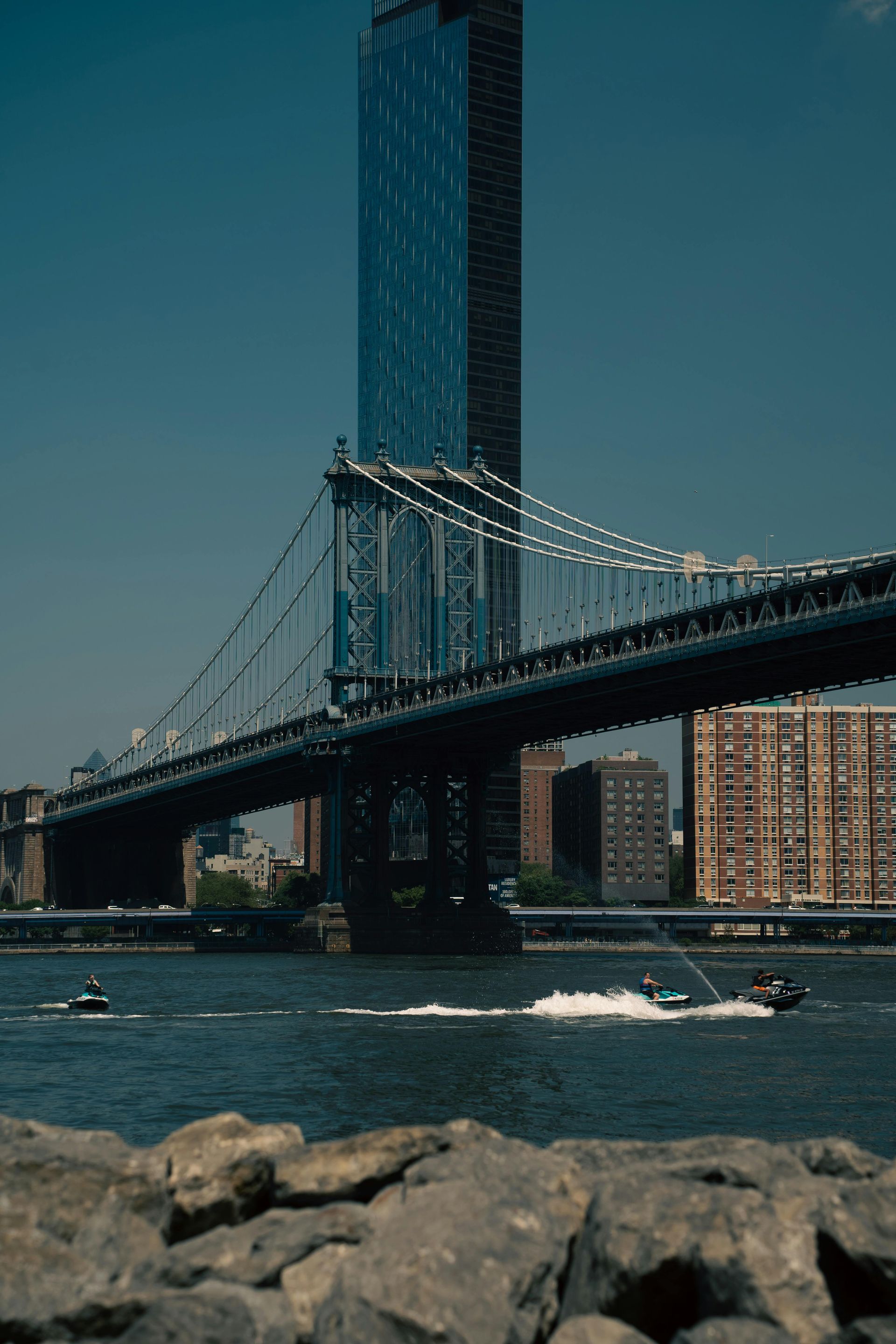 Manhattan Bridge over water, with a tall skyscraper and boats on a sunny day.
