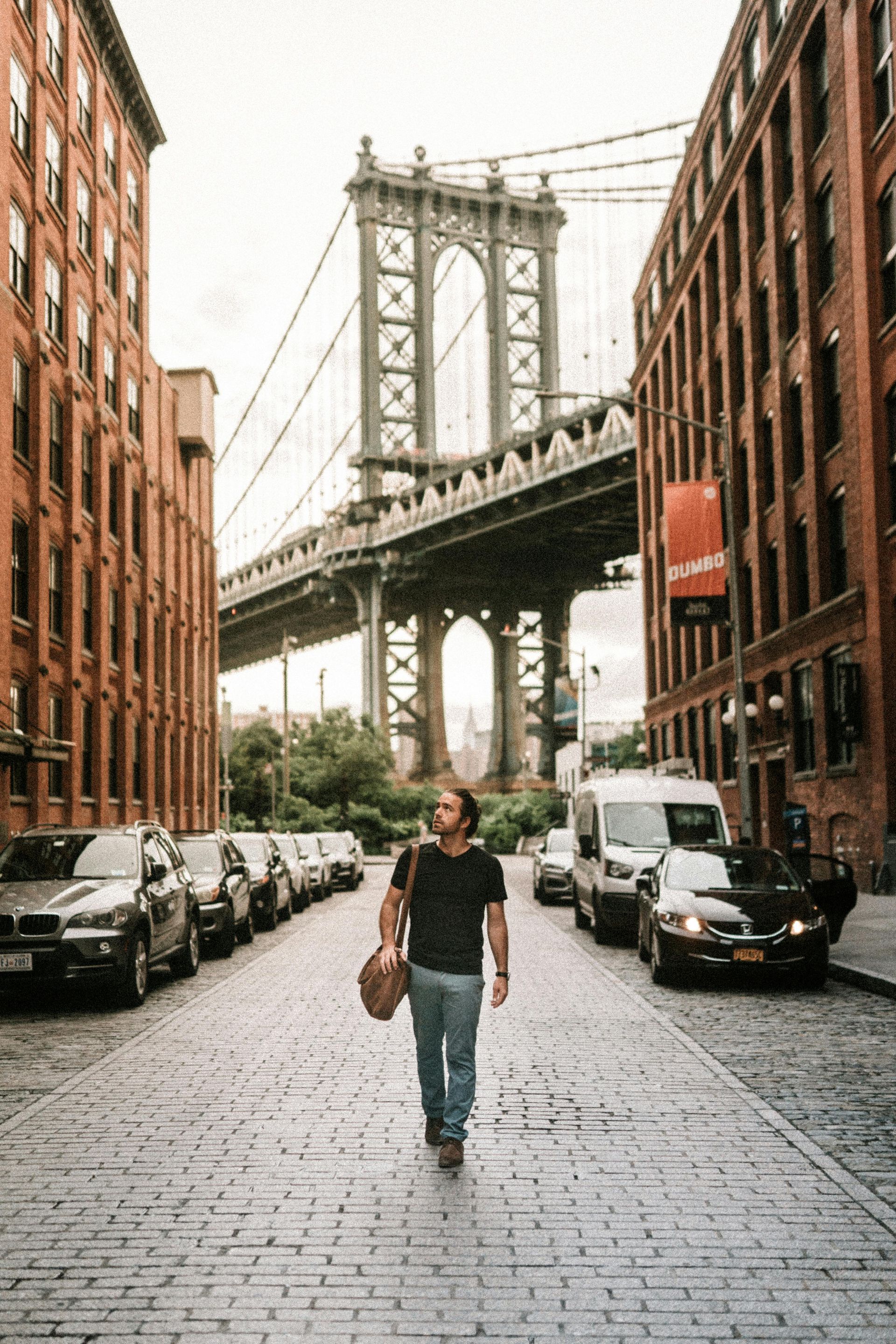 Man walking toward Manhattan Bridge on cobblestone street, brownstone buildings on either side.