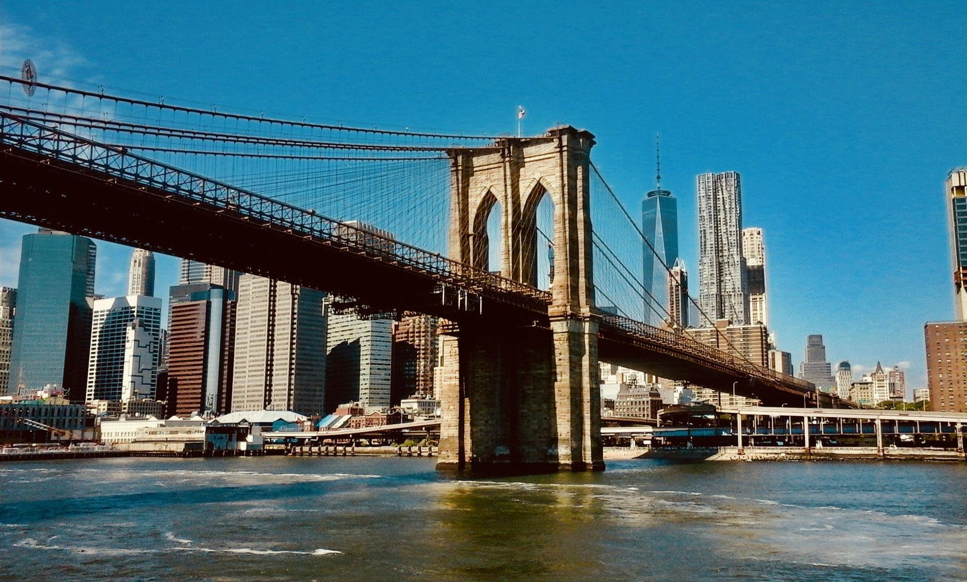 Brooklyn Bridge on a cloudy day, pedestrian walkway with a person taking a photo, city skyline in background.